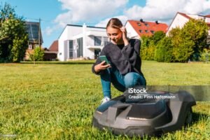 Young woman controlling a robot lawn mower with her smart phone in the summer. The woman kneeling next to the robotic mower, looking to her smart phone to program the robot mower. Modern Home in the background.